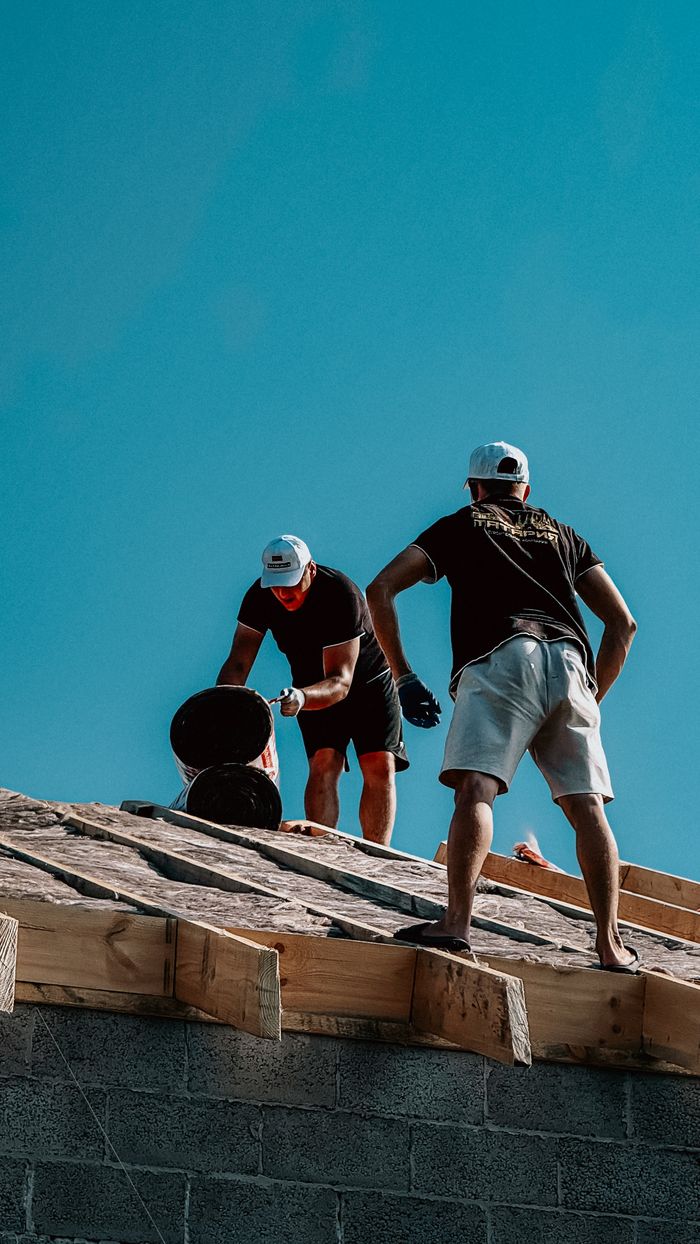 Two construction workers apply roofing materials under a bright blue sky, showcasing teamwork and labor.